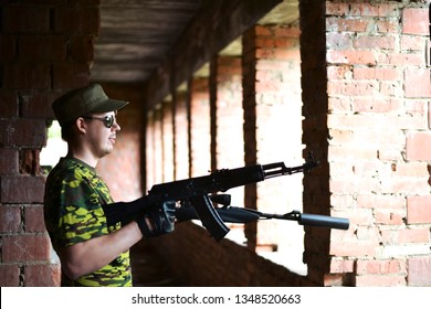 Caucasian Military Man With Black Sunglasses Indoor Urban Room Space Stand With A Kalashnikov Rifle Near Abandoned Red Brick Wall With Windows. Corridor In Perspective. Empty Space For Inscription