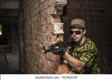 Caucasian Military Man With Black Sunglasses Indoor Urban Room Space Stand With Machine Gun Near Abandoned Red Brick Wall. Dusk Light. Corridor In Perspective.Empty Space For Inscription 