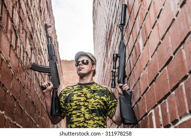 Caucasian Military Man With Black Sunglasses Indoor Urban Room Space Stand With Machine Gun And Rifle With A Telescopic Sight Near Red Brick Wall. Corridor In Perspective.Empty Space For Inscription 