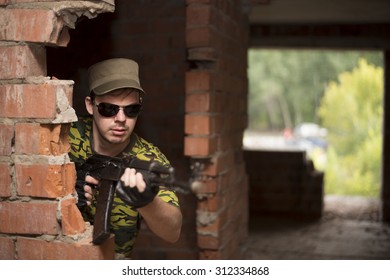 Caucasian Military Man With Black Sunglasses Indoor Urban Room Space Stand With Machine Gun Near Abandoned Red Brick Wall. Dusk Light. Corridor In Perspective.Empty Space For Inscription 