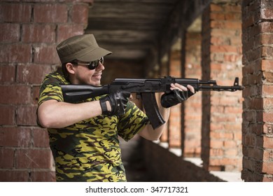 Caucasian Military Man With Black Sunglasses Indoor Urban Room Space Stand With A Kalashnikov Rifle Near Abandoned Red Brick Wall With Windows. Corridor In Perspective.Empty Space For Inscription