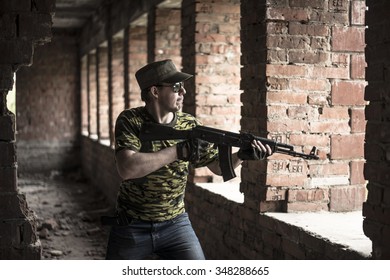 Caucasian Military Man With Black Sunglasses Indoor Urban Room Space Stand With A Kalashnikov Rifle Near Abandoned Red Brick Wall With Windows. Corridor In Perspective.Empty Space For Inscription