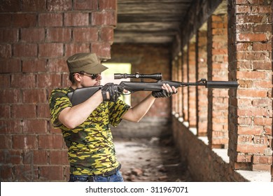 Caucasian Military Man With Sunglasses Indoor Urban Room Space Stand With Rifle With A Telescopic Sight Near Abandoned Red Brick Wall With Windows. Corridor In Perspective.Empty Space For Inscription 