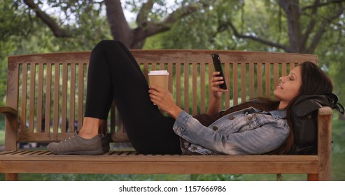 Caucasian Millennial Woman Lying Down On Park Bench And Scrolling Through Phone