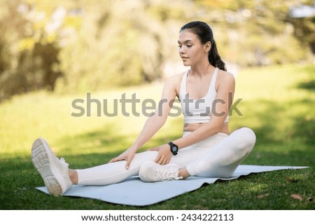 A caucasian millennial woman in white athletic wear gently stretches on a yoga mat in the park, her smartwatch visible as she focuses on her wellness routine, outside, full length