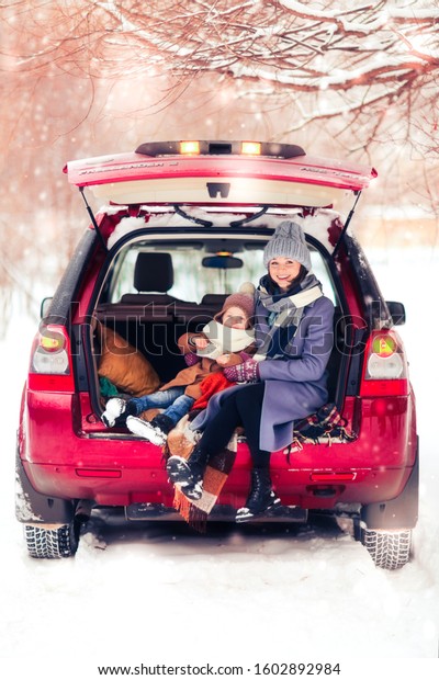 Caucasian mom with daughter child\
with thermos in trunk of car, winter travel with children and\
picnic in car in winter, toning. Magic winter photo with\
snowflakes