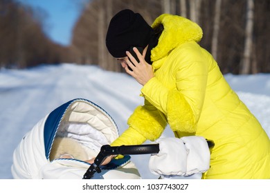Caucasian mom in yellow coat is walking with white baby carriage on snow road among the forest and talking by phone at winter sunny day