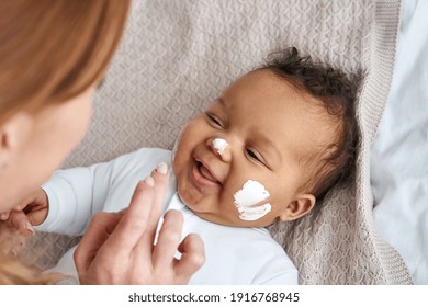 Caucasian mother applying healthy skin care moisturizing cream on cute adorable funny african american baby girl daughter face. Skincare lotion for infant pediatric diathesis treatment concept. - Powered by Shutterstock
