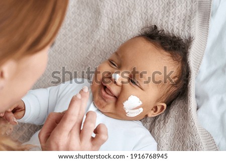 Caucasian mother applying healthy skin care moisturizing cream on cute adorable funny african american baby girl daughter face. Skincare lotion for infant pediatric diathesis treatment concept.