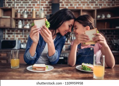 caucasian mother and daughter holding sandwiches and looking at each other in the kitchen