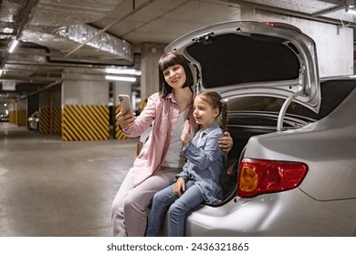 Caucasian mother and preschool daughter making selfie with smartphone in underground parking lot. Caucasian family having video call using cell phone sitting on the trunk of car before exciting trip. - Powered by Shutterstock