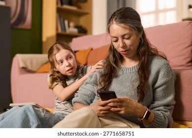 Caucasian mother using smartphone while child daughter touching shoulder at home, showing family disconnection. Useful for parenting, screen time, digital wellbeing campaigns