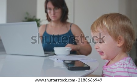 Caucasian Mother Working From Home using Laptop Multitasking Simultaneously Spoon Feeding Baby Girl Daughter Watching Cartoon on Smartphone