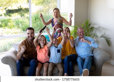 Caucasian multi-generation family in the living room at home, sitting on a couch together, raising hands and cheering while watching TV, the grandson holding an American football - Powered by Shutterstock