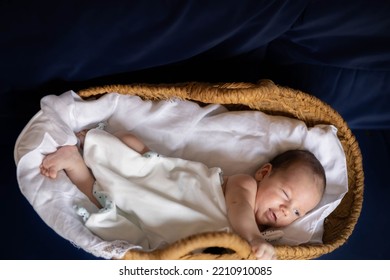 Caucasian Newborn Baby With White Sheets In A Basket And Dark Blurred Background, Making A Funny Wink And A Very Expressive And Tender Expression With His Face