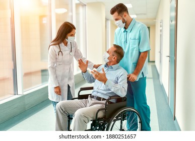 Caucasian Nurse And Handsome Young Doctor Wearing Surgical Masks To Protect Against Covid 19 Pandemic Take Care Of A Mature Male Patient Sitting In A Wheelchair In A Hospital.