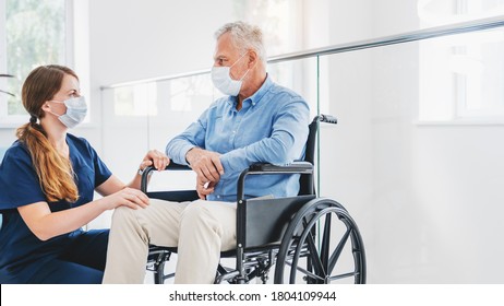 Caucasian Nurse Taking Care Of Senior Male Patient On Wheelchair In Hospital. Young Woman And Old Man Wearing Face Mask For Protection Of Covid 19 Pandemic