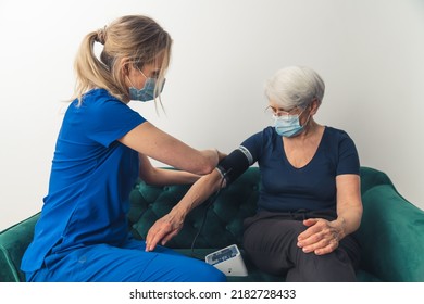 Caucasian Nurse, Wearing A Dark Blue Uniform And A Face Mask, Sitting With Her Senior Woman Patient On A Sofa And Measuring Her Blood Pressure. High-quality Photo