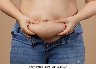 Caucasian obese Woman touching and showing big tummy wearing blue unzipped jeans on beige background. Sudden weight gain. Visceral fat. Body positive. Tight little clothes. Need for wardrobe change.