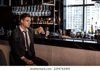 Caucasian office man sitting at bartender counter alone drinking alcohol in rooftop bar after work, lonely young white businessman holding glass of alcohol in nightclub bar, nightlife concept