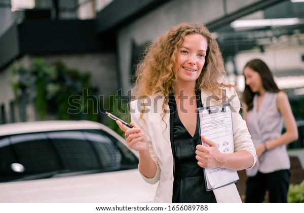 Caucasian officer female staff stands and\
holding rental agreement document in front.  Asian woman holding\
remote car key near vehicle behind in\
background.