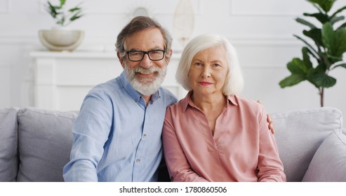 Caucasian Old Gray-haired Married Couple On Pension Sitting On Couch, Looking At Each Other And Then Smiling At Camera. Senior Woman And Man Resting On Sofa At Home. Portrait Shot.