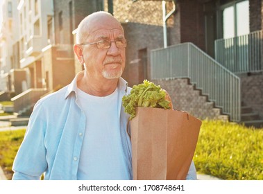 Caucasian Old Man In Glasses Walking The Street Home After Shopping And Carrying Packet With Grocery. Male Senior Retired Pensioner Holding And Carrying Food In Bag Outdoors. Coming Back From Market.