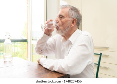 Caucasian old man with white beard and glasses drinks a glass of fresh water in the kitchen - Powered by Shutterstock