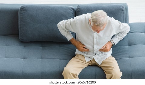 Caucasian Old Senior Elderly Stressed Unhealthy Gray Hair And Bearded Man Sitting On Sofa In Living Room At Home Alone Holding Hands On Belly Suffering From Painful Indigestion Diarrhea Stomachache.