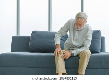 Caucasian Old Senior Elderly Unhealthy Sick Ill Grey Bearded Male Husband Grandpa Sitting On Sofa At Home Alone Holding Hands On Knee Having Emergency Painful Joint And Muscle Ache Injury Problem.