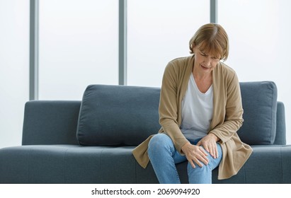 Caucasian Old Senior Elderly Unhealthy Sick Ill Female Housewife Grandmother Sitting On Sofa At Home Alone Holding Hands On Knee Suffering From Emergency Painful Ache On Muscle And Joint Problem.