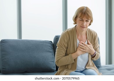 Caucasian Old Senior Elderly Unhealthy Sick Ill Female Housewife Sitting On Sofa At Home Alone Holding Hands On Chest Having Emergency Cardio Problem Suffering From Urgent Heart Attack Need Ambulance.