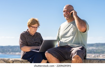 Caucasian Older Woman Sitting Using A Laptop Next To A Smiling Middle-aged Man Talking On The Phone, On A Sunny Day