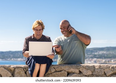 Caucasian Older Woman Sitting Using A Laptop Next To A Smiling Middle-aged Man Talking On The Phone, On A Sunny Day