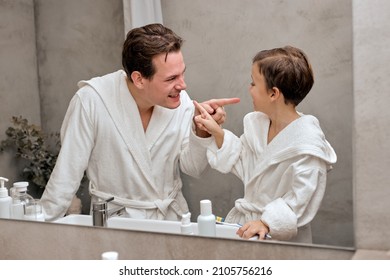 Caucasian Parent Man With Child Boy In White Bathrobes Having Morning Routine In Hotel Bathroom, Standing Near The Mirror. Dad With Son Examining Teeth. Family Vacation, Healthcare Concept.