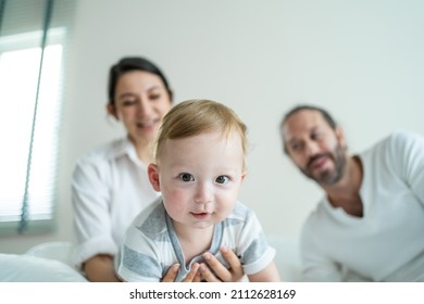 Caucasian parents play with cute baby boy child infant on bed at home. Happy family, attractive mom and dad look to toddler son crawl with happiness enjoy after wake up activity in bedroom in house.