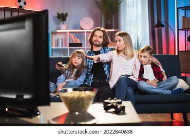 Caucasian Parents With Son And Daughter Sitting In Embrace On Sofa And Watching Movie During Evening Time. Young Family In Casual Wear Spending Time Together At Home.