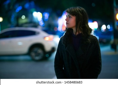 Caucasian pedestrian woman walking the street at night in the city with moving cars in the background.  She looks nervous or unsure like a lost tourist or afraid of commuting alone. 