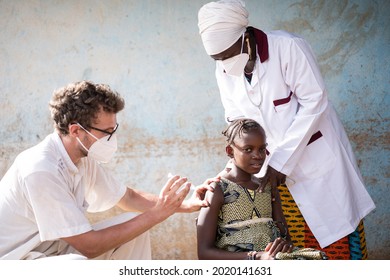 Caucasian pediatrician doctor is vaccinating a brave little African girl supported by a kind black nurse who is holding her sholders. Both healthcare workers wear protective mask to prevent from catch