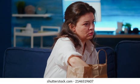 Caucasian Person Removing Face Mask After Getting Takeout Meal In Delivery Bag On Table At Home. Young Woman Preparing Food For Eating While Watching Television In Living Room After Work