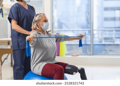 Caucasian physiotherapist and senior woman with artificial leg with face masks using exercise band. Hospital, hygiene, disability, physiotherapy, work, medicine and healthcare, unaltered. - Powered by Shutterstock