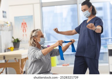 Caucasian physiotherapist and senior woman with face masks using exercise band stretching arms. Hospital, hygiene, physiotherapy, work, medicine and healthcare, unaltered. - Powered by Shutterstock