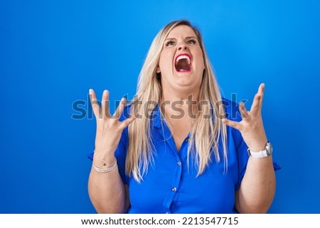 Caucasian plus size woman standing over blue background crazy and mad shouting and yelling with aggressive expression and arms raised. frustration concept. 