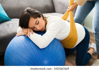 Caucasian Pregnant Woman Doing Breathing Exercises And Resting On A Fitness Ball While Her Doula Wraps A Rebozo Around Her Round Belly