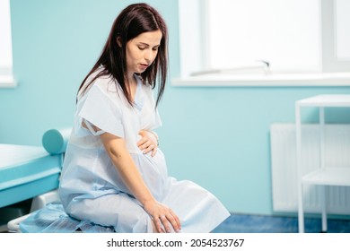 Caucasian Pregnant Woman With Painful Contraction Sitting At Hospital Ward, Touching Belly And Looking Down. Childbirth Process At Hospital.