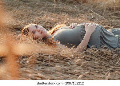 Caucasian pregnant young red-haired woman in dress lying on wheat field in summer at sunset, future mother relaxing in nature, healthy and happy pregnancy concept, new life