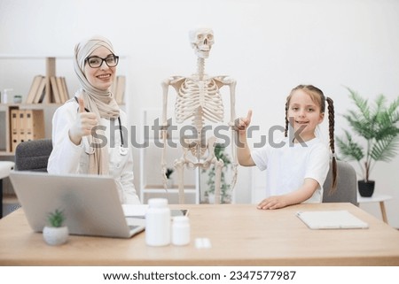 Caucasian preteen girl in casual wear touching sternum on human teaching model at doctor's workplace in clinic showing thumbs up