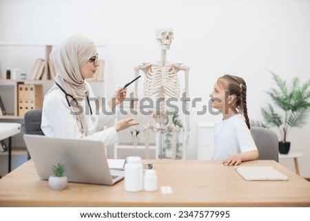 Caucasian preteen girl in casual wear touching sternum on human teaching model at doctor's workplace in clinic. Muslim physician in headscarf explaining importance of breastbone in body functioning.