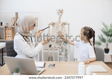 Caucasian preteen girl in casual wear touching sternum on human teaching model at doctor's workplace in clinic. Muslim physician in headscarf explaining importance of breastbone in body functioning.