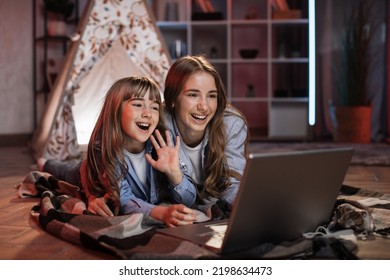 Caucasian Pretty Blond Sisters Lying On Floor Outside Wigwam While Using Laptop For Watching Movie During Evening Leisure Time. Cozy Home And Family Concept.